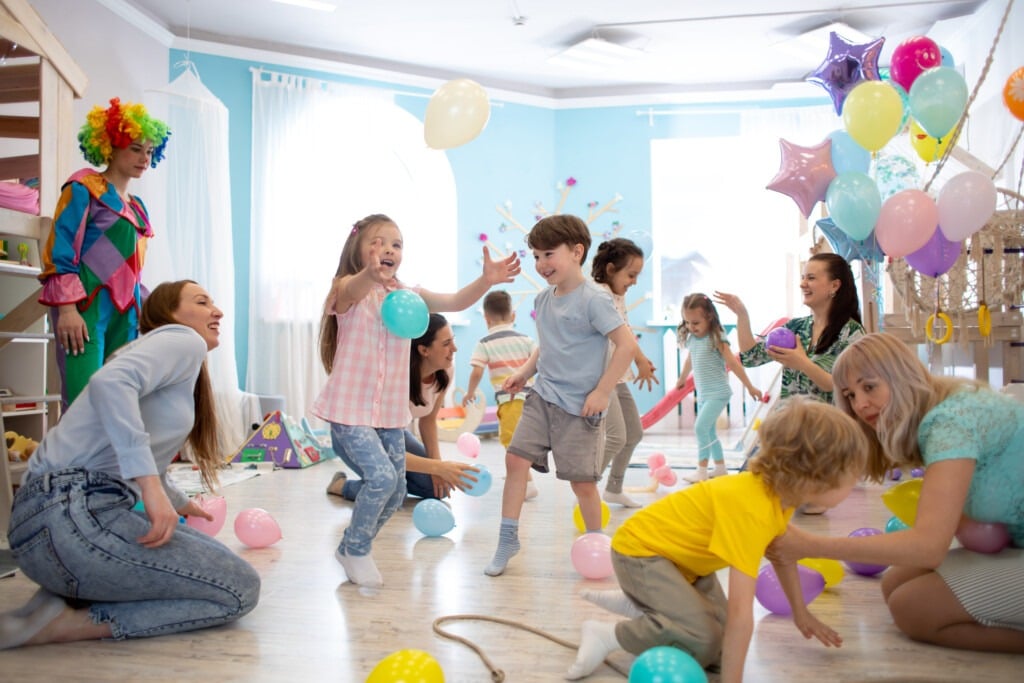 A group of children play with balloons in a bright, colorful room while adults supervise, reflecting the joyful atmosphere at autism services near Houston TX. A clown in the background adds to the lively and festive scene.