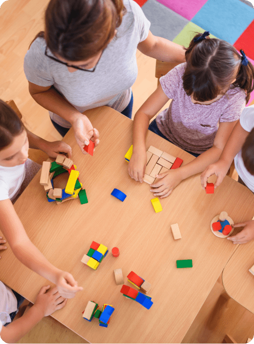 Four children and an adult gather around a wooden table, playing with colorful building blocks. The lively scene, set on a vibrant rug, reflects the engaging environment of ABA near Houston TX.