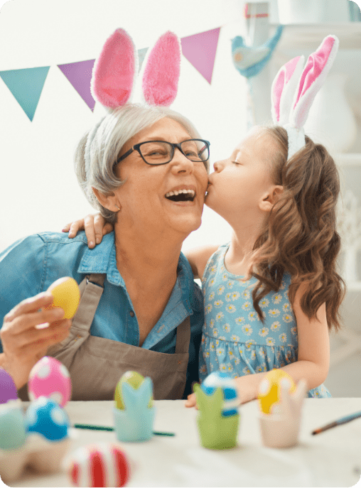 A smiling older woman and a young girl wearing bunny ears decorate Easter eggs together; the girl kisses the woman’s cheek in a festive, colorful room with bunting—an uplifting moment that highlights connections fostered by ABA near Katy TX.