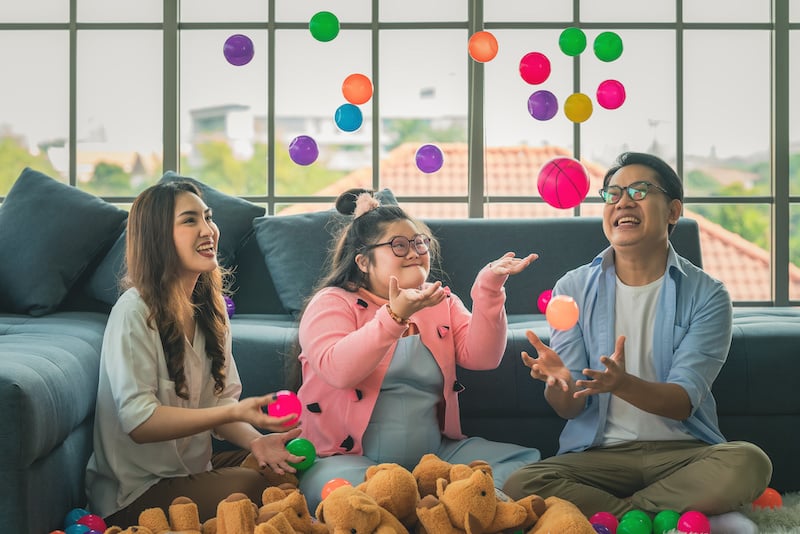 Three people sit indoors on the floor, smiling and tossing colorful plastic balls in the air, enjoying playful moments often seen during autism therapy near Houston TX, with stuffed animals scattered nearby and a large window in the background.