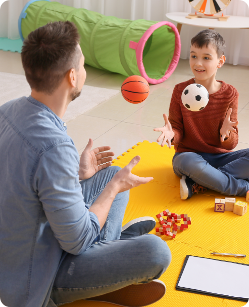 An adult and a child sit on a yellow mat indoors, smiling and tossing a basketball and soccer ball to each other during an ABA near Houston TX session. Toys, blocks, a clipboard, and a green tunnel are visible in the background.