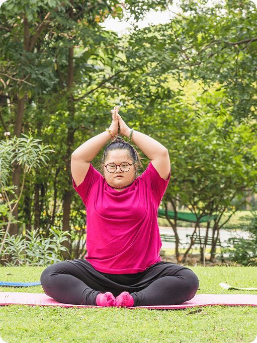 A person sits cross-legged on a yoga mat outdoors, wearing a bright pink shirt and glasses, with their hands pressed together above their head in a yoga pose—perfect for anyone seeking calm, including those looking for an autism therapist near Houston TX.