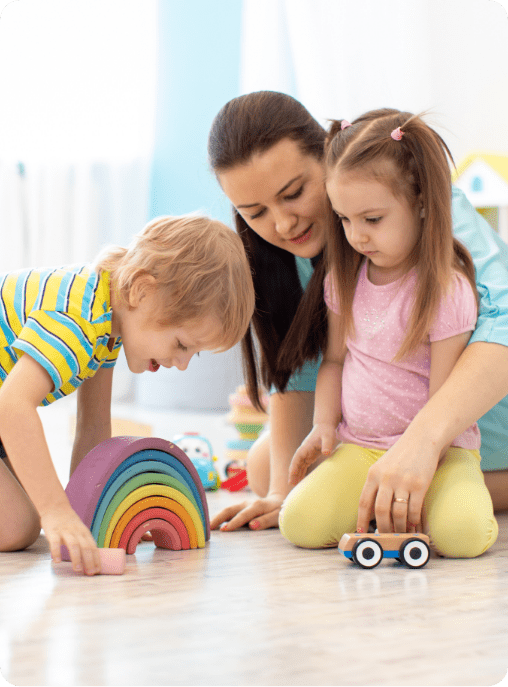 A woman and two young children play together on the floor. The boy enjoys a rainbow stacking toy while the girl touches a small car. All three look focused and happy, illustrating the benefits of autism therapy near Houston TX.