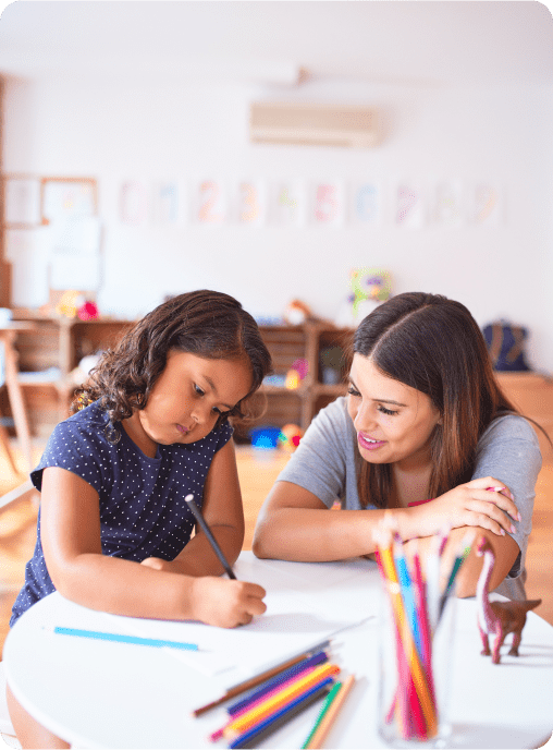 A young girl writes or draws at a table while a woman, possibly her teacher or parent, smiles supportively. Colored pencils and a toy dinosaur sit nearby, reflecting the welcoming environment of ABA near Houston TX. A classroom is in the background.