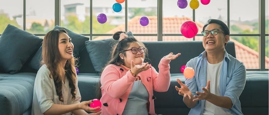 Three people sit on the floor in a living room, smiling and tossing colorful plastic balls in the air. A large window and a couch are in the background. The atmosphere is joyful and playful.
