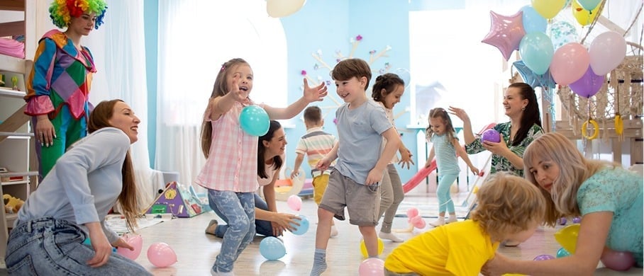 Children and adults are playing with balloons at a lively indoor party. A person dressed as a clown stands in the background. The room is decorated with pastel colors and filled with laughter and movement.