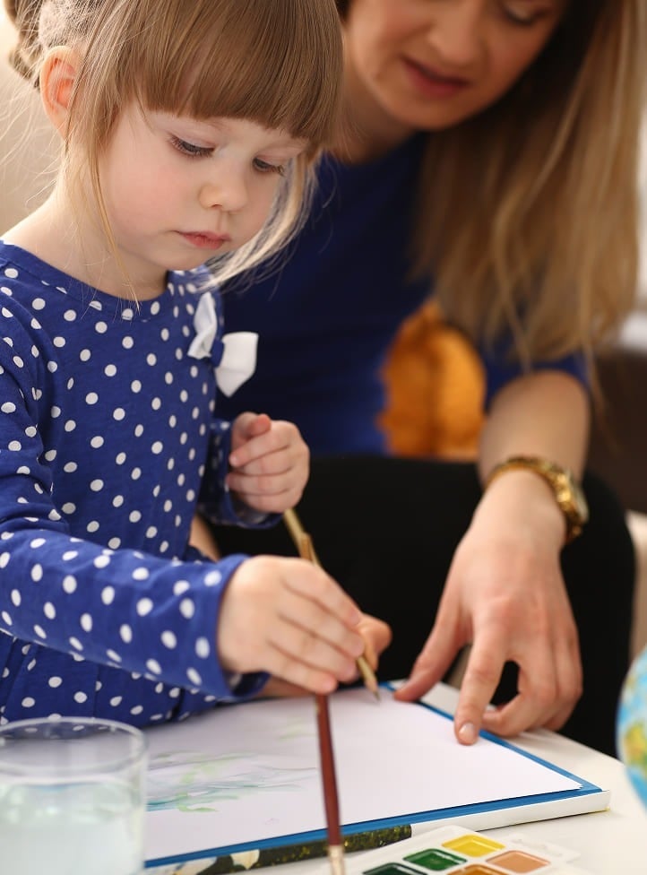 A young girl in a blue polka dot shirt paints on paper with a brush, while an adult woman sits beside her, gently guiding her hand. A palette and glass of water are on the table.