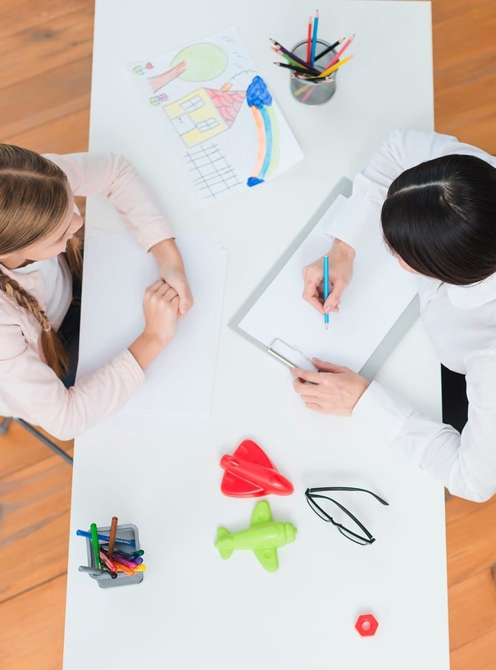 Two people sit across from each other at a white table with drawing supplies, a child’s drawing, colorful toy airplanes, a pencil holder, and glasses. One person is writing while the other watches.