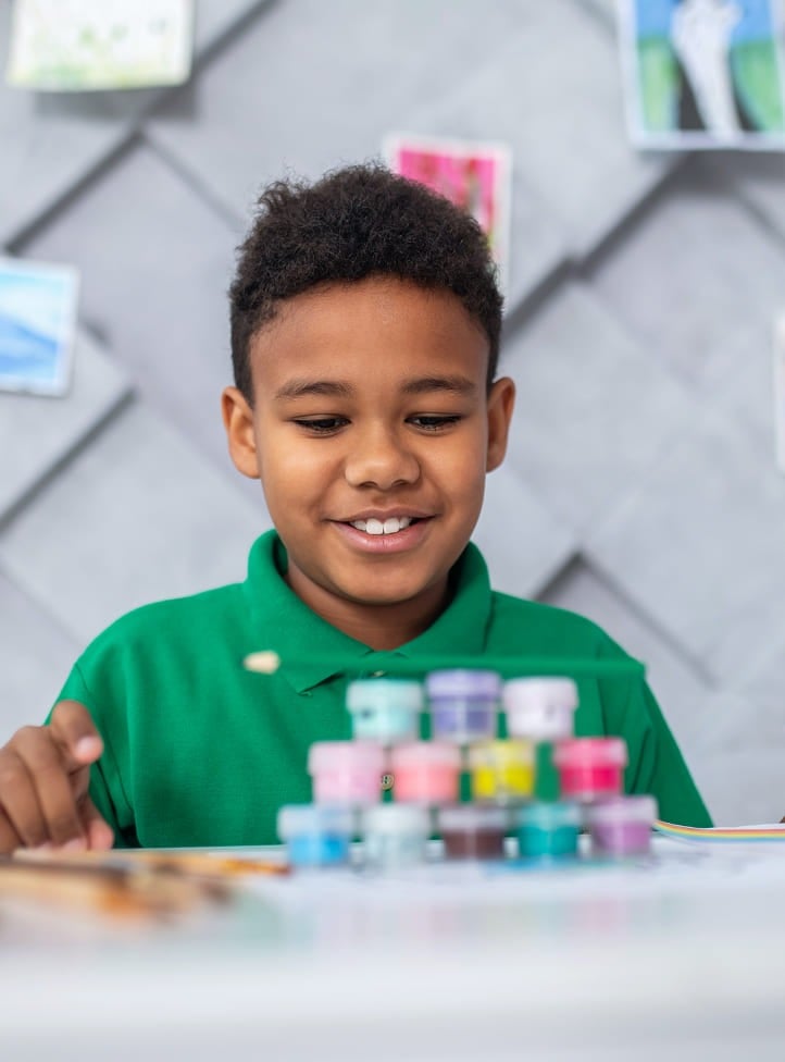 A young boy in a green shirt smiles as he looks at a colorful stack of small paint containers on a table, with art supplies and artwork visible in the background.