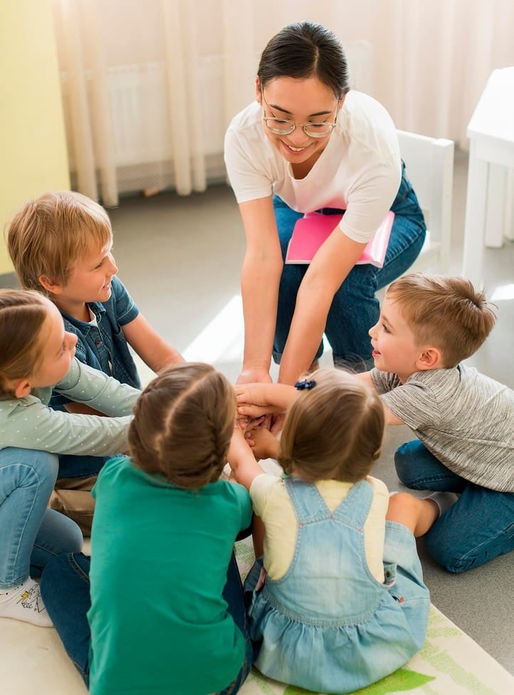 A smiling adult sits on the floor with five young children, all placing their hands together in the center, suggesting teamwork or a group activity in a bright, cheerful room.
