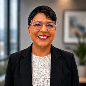 Smiling person with short dark hair, wearing clear glasses, a black blazer, and a white patterned shirt, standing in a modern office with blurred artwork and a plant in the background.