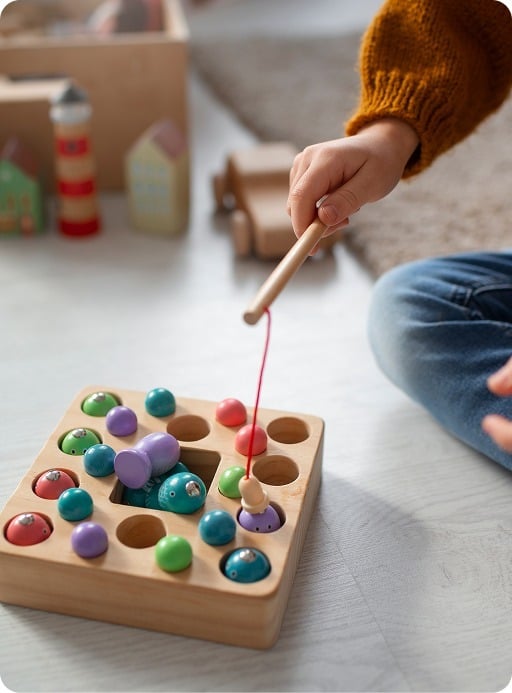 A child wearing a brown sweater uses a wooden fishing rod toy to pick up colorful, round pieces from a wooden board game on the floor.