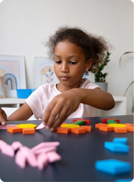 A young girl with curly hair sits at a table, concentrating as she arranges colorful geometric shapes. There are framed pictures and a plant in the background.