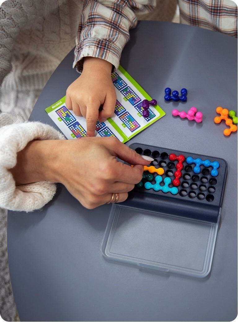 Two people play a logic puzzle game with colorful pieces on a black grid board. One hand points at a guide sheet, while the other places an orange piece on the board. The scene is on a round gray table.