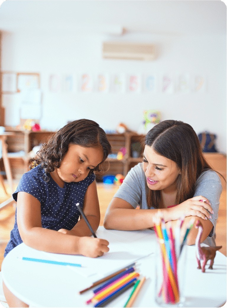 A young girl writes or draws on paper at a table while an adult woman sits beside her, smiling and watching. Colored pencils are in the foreground, and a classroom setting is visible in the background.